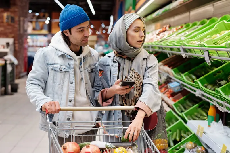 Un couple fait l'épicerie avec un panier ; elle consulte son téléphone devant l'étalage de légumes.