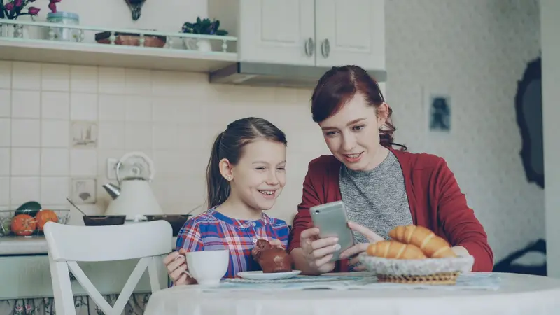Une mère et sa fille regardent un téléphone en souriant, attablées dans la cuisine au déjeuner.
